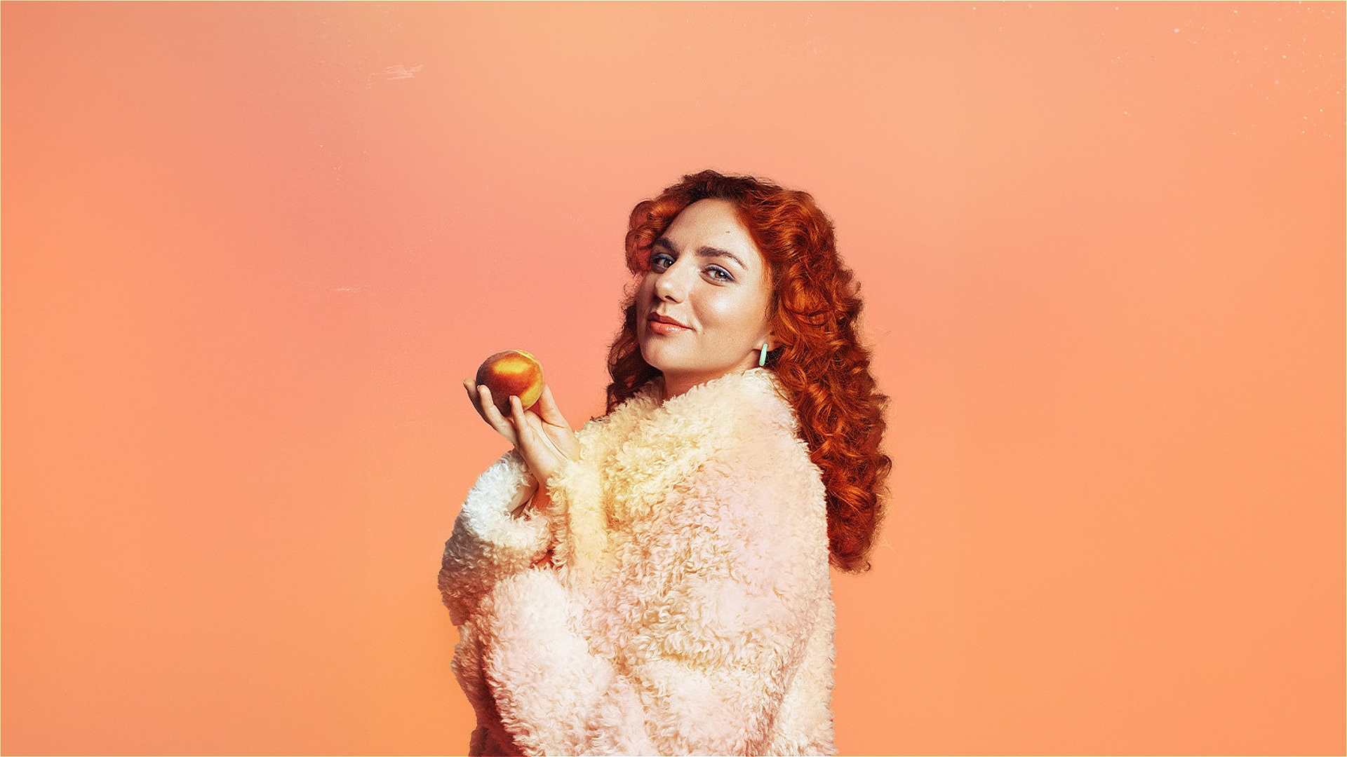 Comedian Ania Magliano holding a peach in a fluffy coat, against an orange background