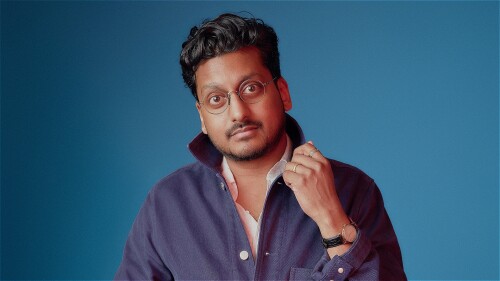 The comedian Ahir Shah stands in front of a blue background, wearing a navy shacket over a white shirt.