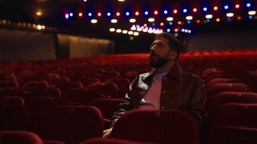 A man with a beard sits in an empty auditorium surrounded by red chairs, with shiny lights blurred in the background