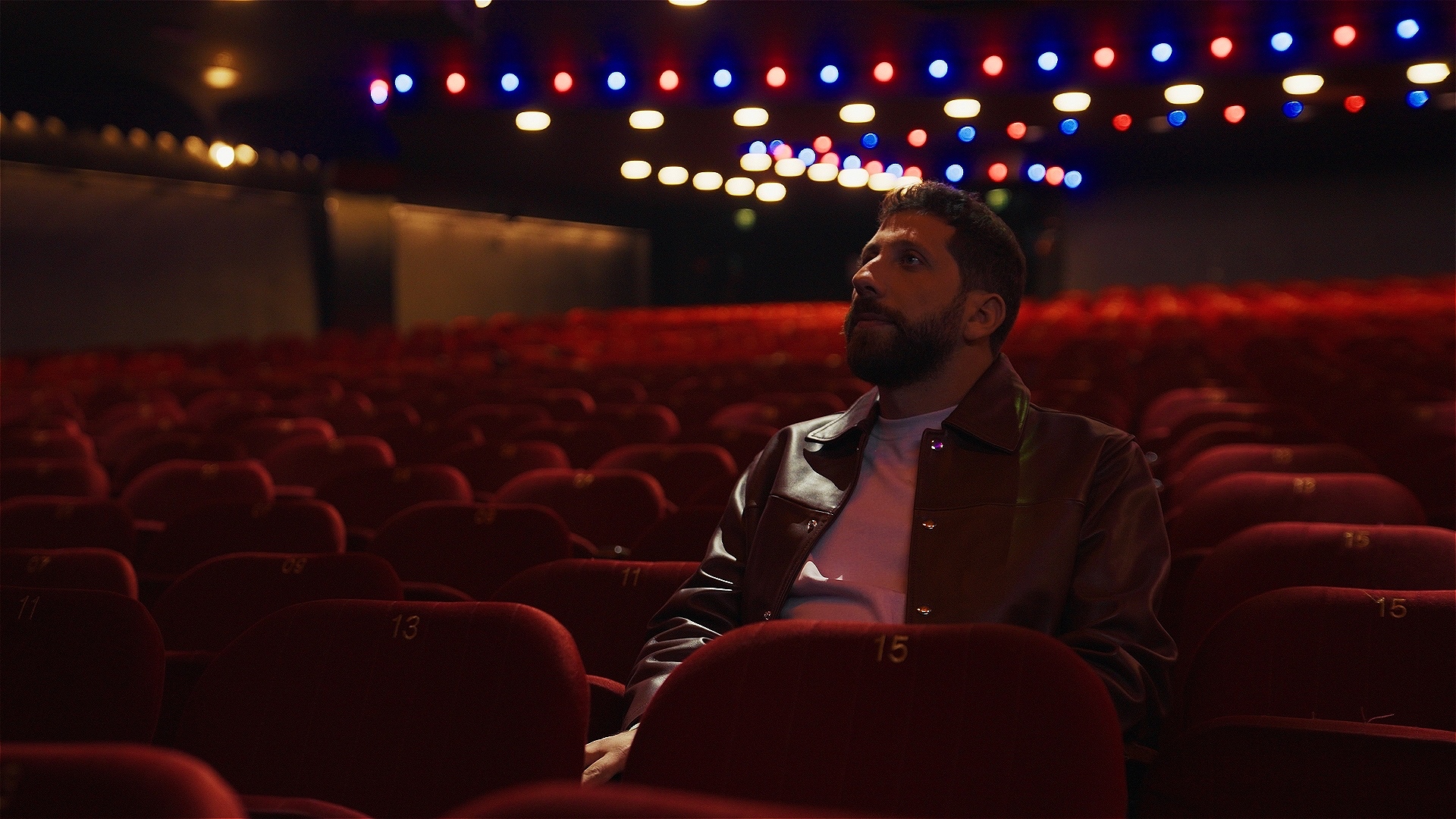 A man with a beard sits in an empty auditorium surrounded by red chairs, with shiny lights blurred in the background